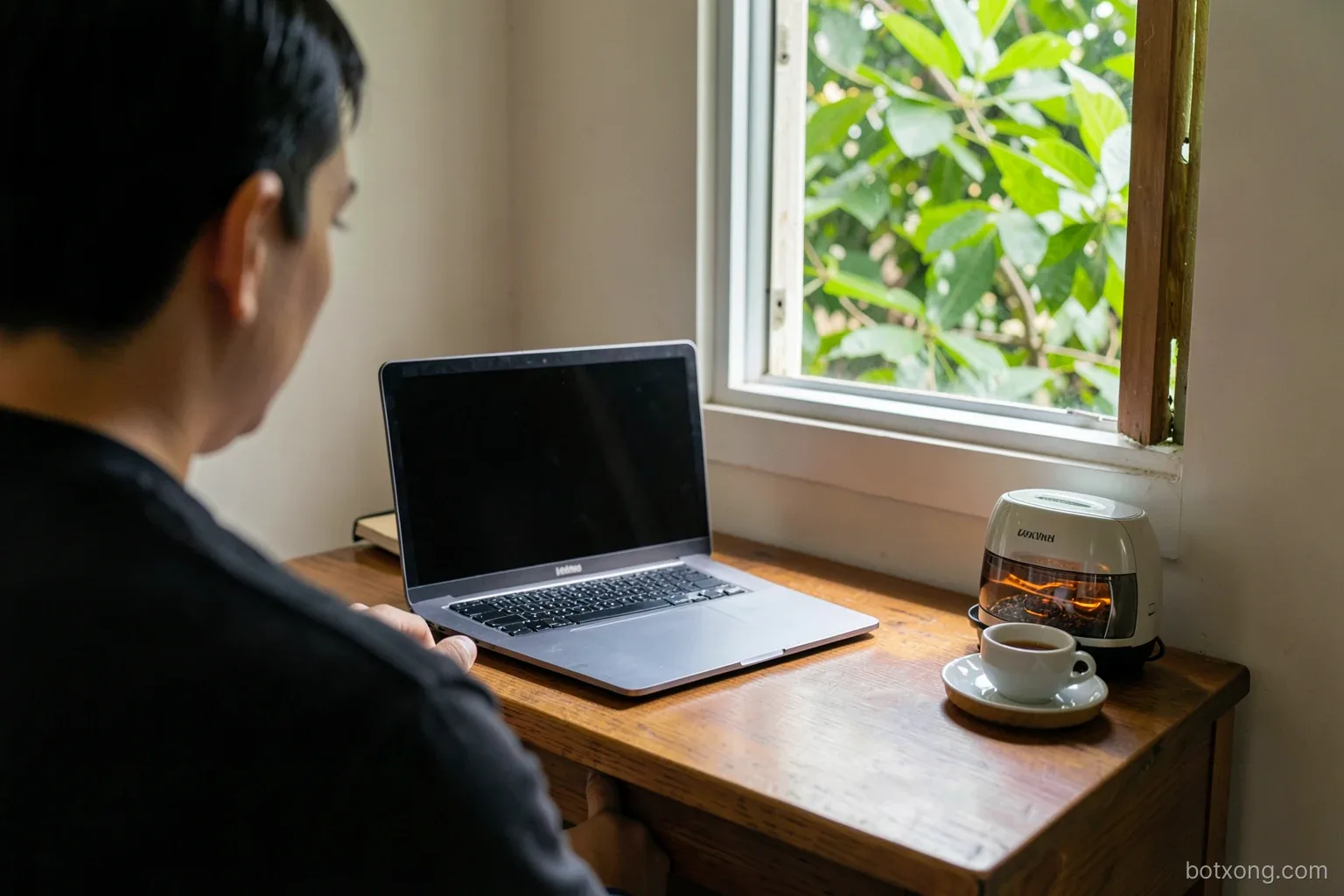 Over-the-shoulder view of a home study corner in Vietnam, wooden desk with a closed laptop and teacup, small electric agarwood warmer on a side shelf, window half open to green leaves